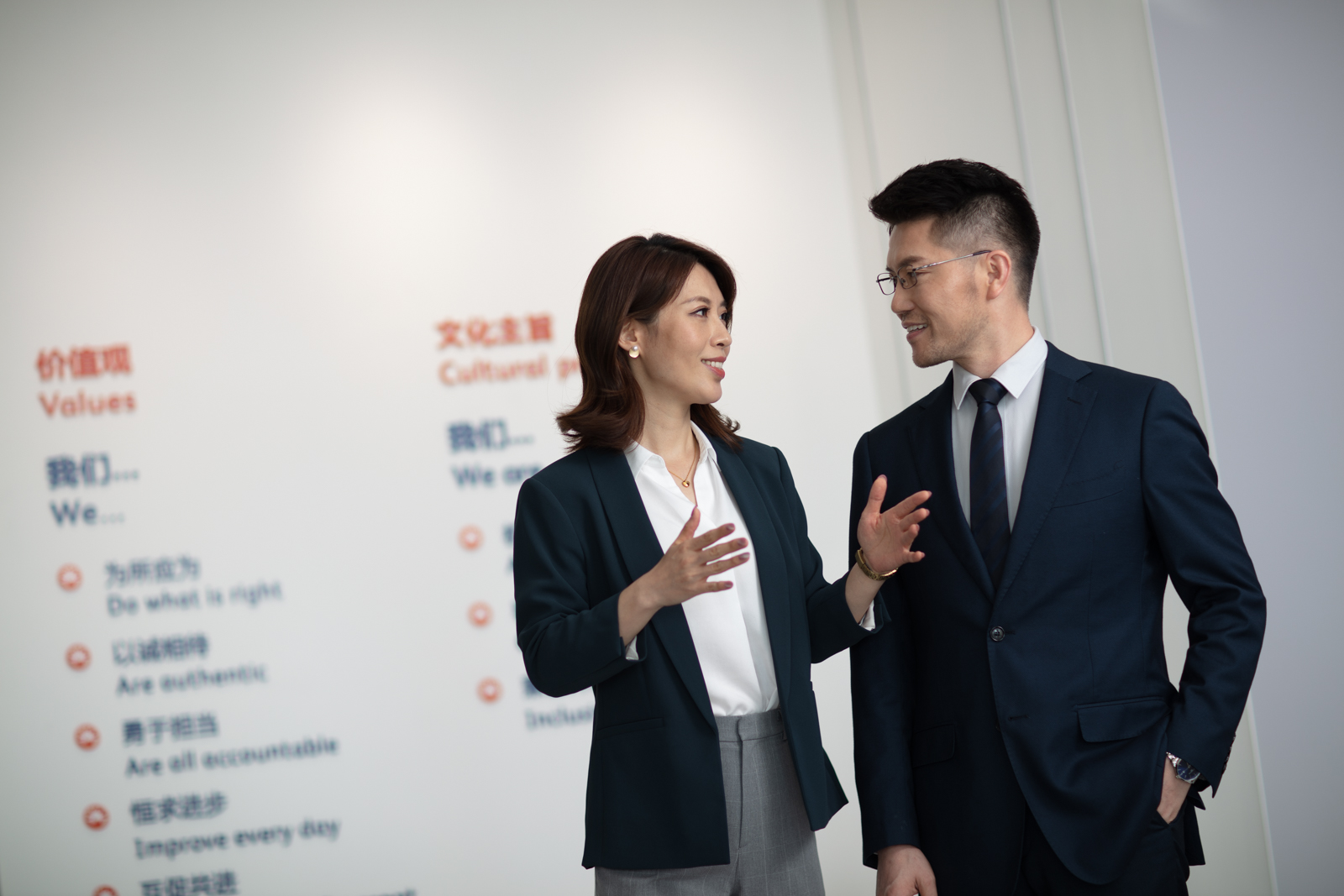 Two smiling business professionals in formal attire speaking in a bright office with bilingual BD values on the wall